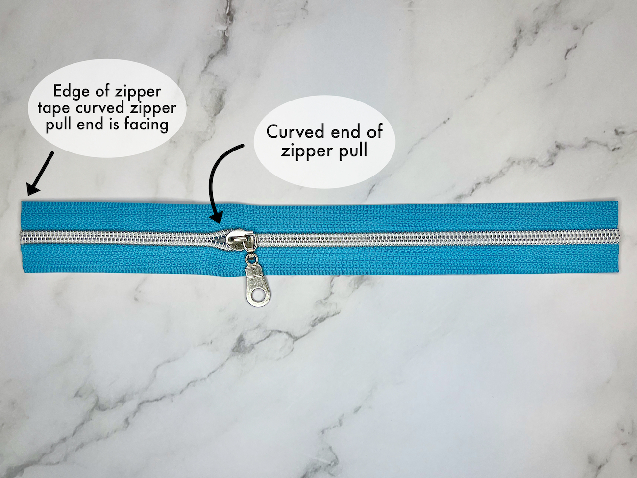 Zipper tape with zipper pull with an arrow pointing to one edge of the zipper tape and one arrow pointing to the curved edge of the zipper pull