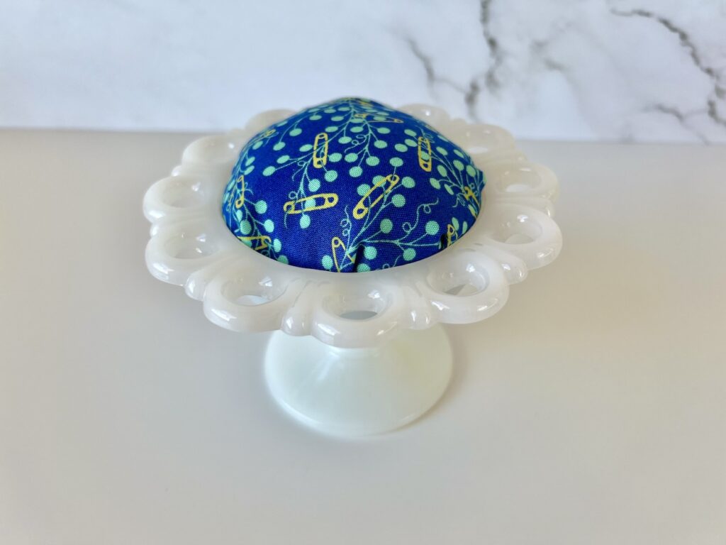 A white, milk glass candy dish with a blue fabric pincushion top