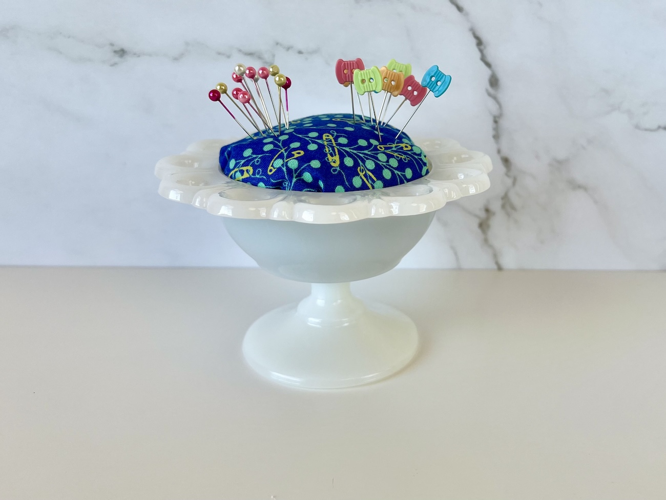 A white, milk glass candy dish with a blue fabric pincushion top with colorful pins in the pincushion