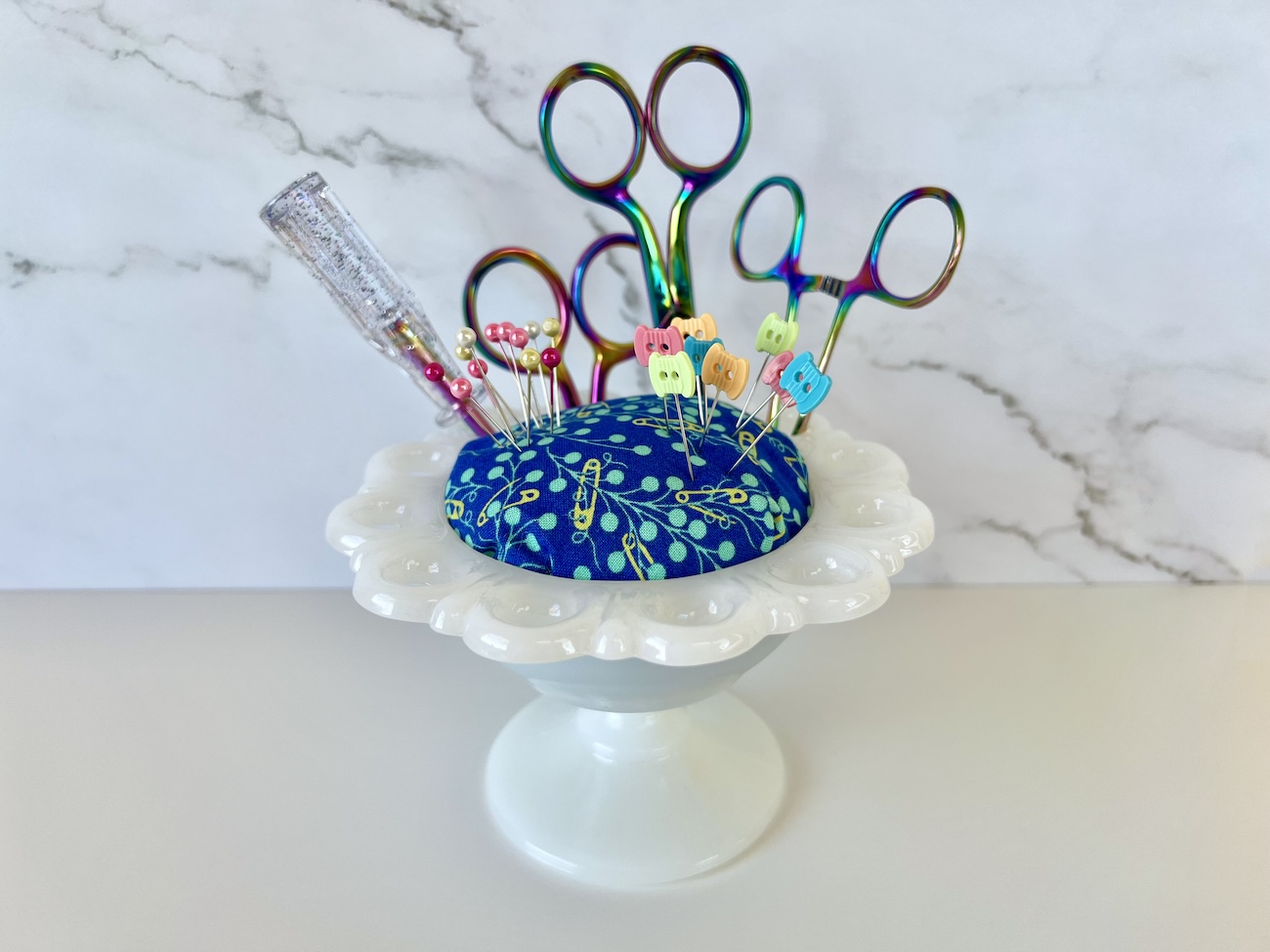 A white, milk glass candy dish with a blue fabric pincushion top with colorful pins in the pincushion, and rainbow scissors in the candy dish